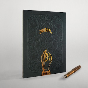 May include: A black journal with a gold embossed floral design and the word "JOURNAL" on the cover. A gold hand with a pen is also embossed on the cover. A gold fountain pen is lying on a white surface.