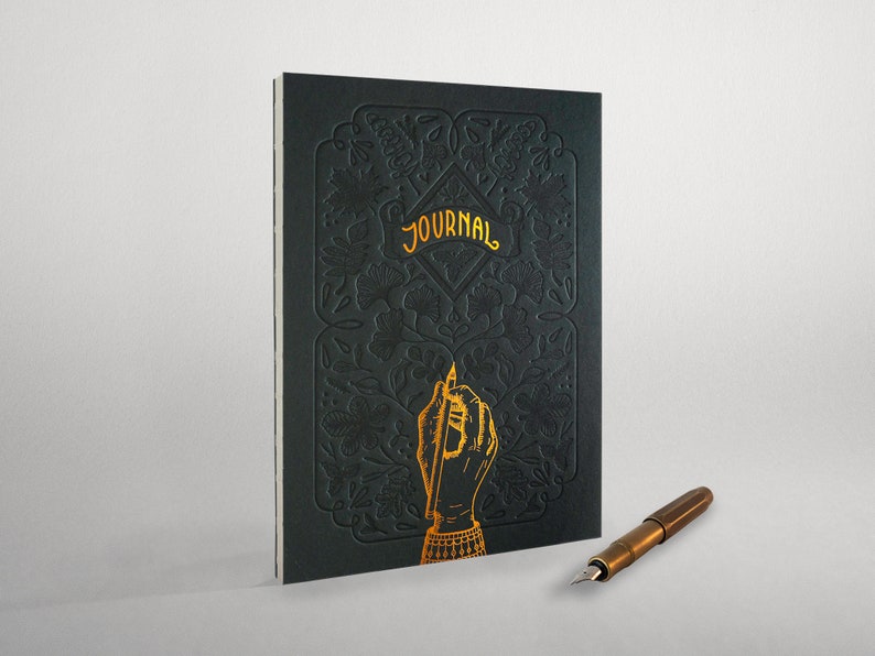 May include: A black journal with a gold embossed floral design and the word "JOURNAL" on the cover. A gold hand with a pen is also embossed on the cover. A gold fountain pen is lying on a white surface.