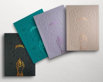 May include: Four different colored journals with embossed floral designs and the word "JOURNAL" in gold lettering. Each journal features a hand holding a pen.