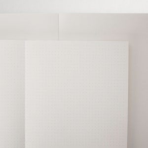 May include: A white notebook with a dotted grid pattern, open to a blank page. The notebook is on top of a white surface.