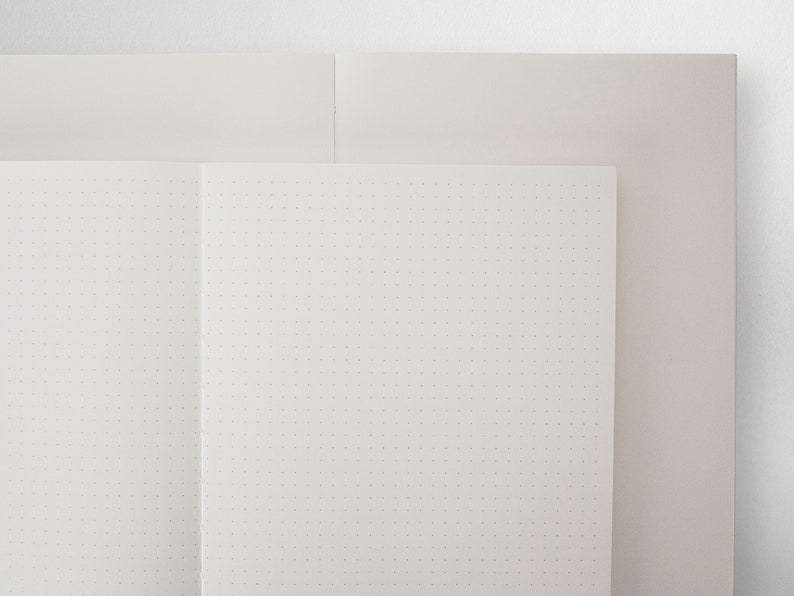May include: A white notebook with a dotted grid pattern, open to a blank page. The notebook is on top of a white surface.