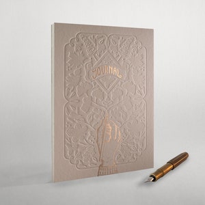 May include: A beige journal with a gold embossed floral design and the word "JOURNAL" in gold lettering. A gold fountain pen is lying on a white surface.