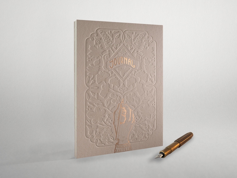May include: A beige journal with a gold embossed floral design and the word "JOURNAL" in gold lettering. A gold fountain pen is lying on a white surface.
