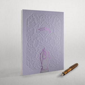 May include: A purple journal with an intricate floral design and the word "JOURNAL" embossed on the cover. A hand holding a pen is depicted on the front cover. A gold fountain pen is lying on a white surface.
