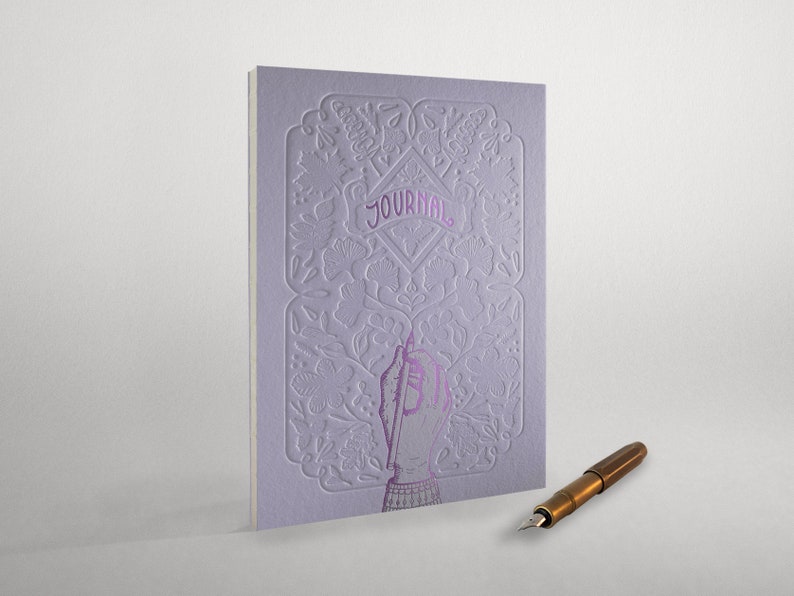 May include: A purple journal with an intricate floral design and the word "JOURNAL" embossed on the cover. A hand holding a pen is depicted on the front cover. A gold fountain pen is lying on a white surface.