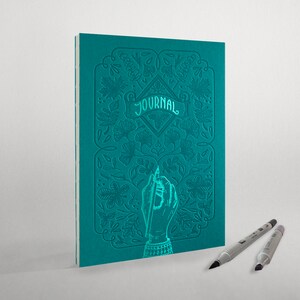 May include: Teal blue journal with an embossed floral design and the word "JOURNAL" on the cover. A hand holding a pen is also embossed on the cover. Two silver pens are lying on a white surface.