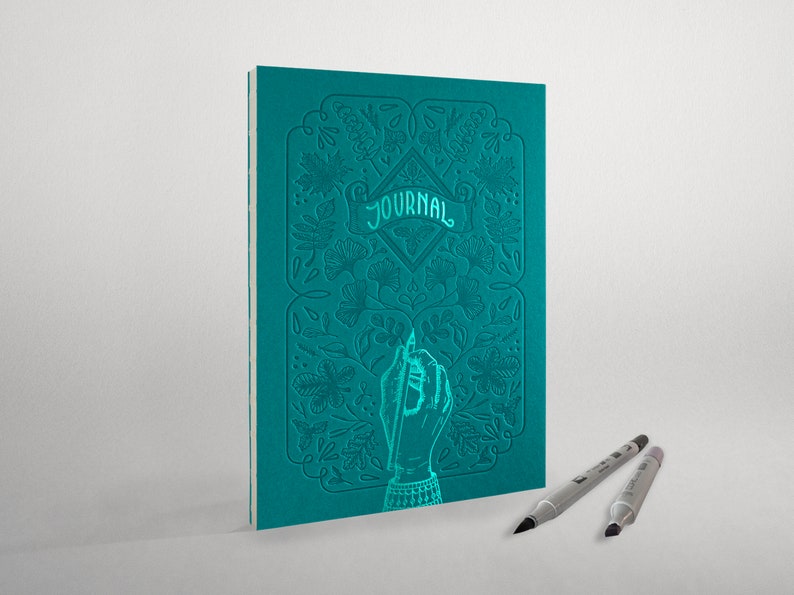 May include: Teal blue journal with an embossed floral design and the word "JOURNAL" on the cover. A hand holding a pen is also embossed on the cover. Two silver pens are lying on a white surface.