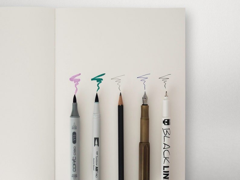 May include: A close-up of six writing instruments: a grey Copic Ciao marker, a green Tombow ABT dual brush pen, a black pencil, a gold fountain pen, a white Black Line pen, and a white pen with a black cap.