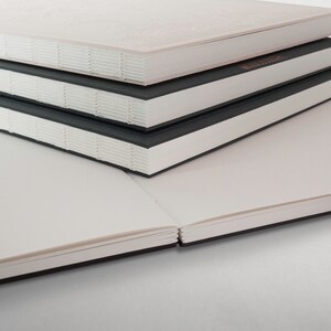 May include: A stack of four blank notebooks with black covers and white pages. The notebooks are closed and stacked on top of each other.