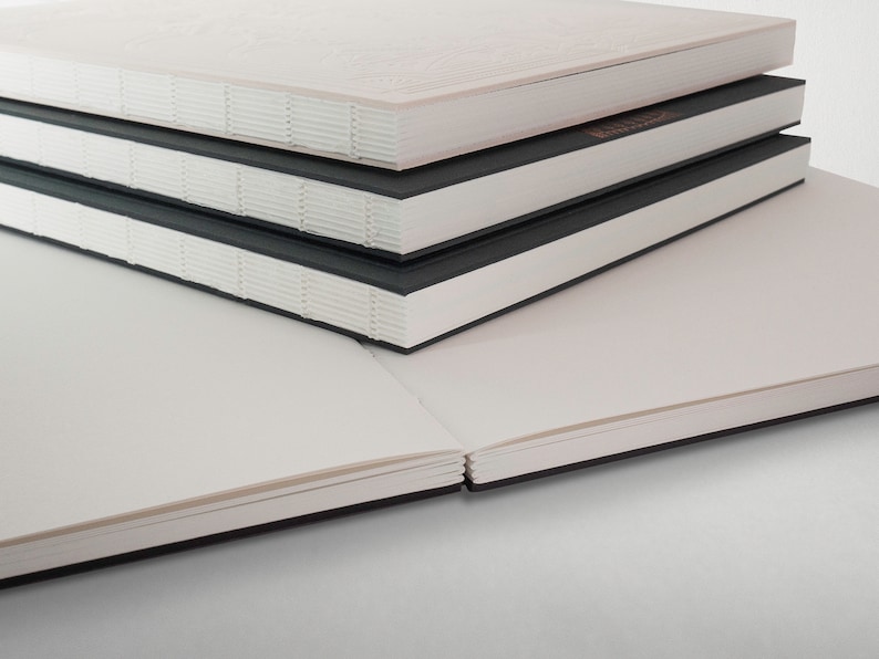 May include: A stack of four blank notebooks with black covers and white pages. The notebooks are closed and stacked on top of each other.