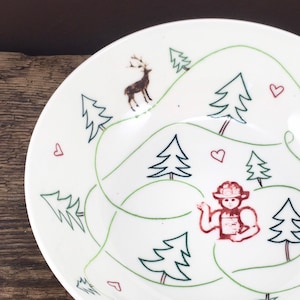 Puede incluir: Tazón de cerámica blanco con un diseño verde y rojo de una escena de bosque. El diseño incluye árboles, un ciervo, un corazón y una persona con un sombrero rojo.