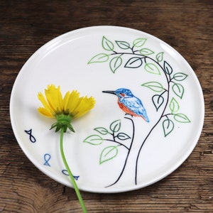 Op de afbeelding: Witte keramische plaat met een handgeschilderd ontwerp van een blauw-oranje ijsvogel op een tak met groene bladeren. Een gele bloem ligt op de plaat.