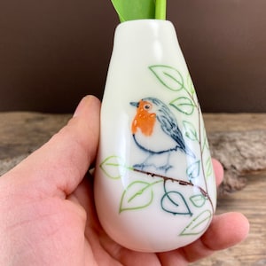 Può includere: Un vaso a bocciolo in ceramica bianca con un'illustrazione dipinta a mano di un pettirosso posato su un ramo con foglie verdi. L'uccello ha il petto arancione, le ali grigie e un becco nero. Il vaso contiene uno stelo verde.
