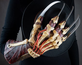 May include: A red and white Freddy Krueger glove with long, sharp blades. The glove is worn by a person wearing a black shirt and a black hat.