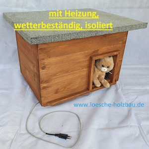 Peut inclure: Une maison pour chat en bois avec un toit gris et un chat en peluche qui sort de l'entrée. La maison est équipée d'un cordon, indiquant qu'elle possède un élément chauffant. Le texte "mit Heizung, wetterbeständig, isoliert" et "www.loesche-holzbau.de" est imprimé sur le toit.
