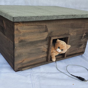 Peut inclure: Une maison pour chat en bois avec un toit en feutre gris et une petite entrée. Un jouet en peluche pour chat sort de l'entrée. Un cordon électrique gris est connecté à la maison.