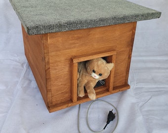 Può includere: Una casetta per gatti in legno con tetto grigio e apertura rettangolare. Un gatto di peluche sbuca dall'apertura. La casetta per gatti ha un cavo di alimentazione con spina.