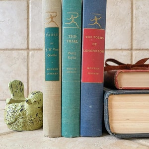 May include: Three vintage hardcover books with gold lettering on the spines. The books are titled "Faust", "The Trial", and "The Poems of Longfellow". The books are arranged vertically, with the tallest book on the left and the shortest book on the right. The books are sitting on a light brown tiled surface.