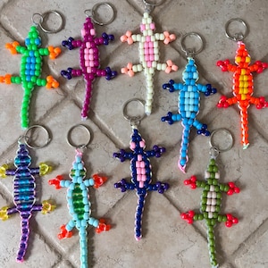 May include: A collection of nine lizard-shaped keychains crafted from colorful plastic pony beads. Each lizard features a distinct color combination, including green, purple, pink, blue, orange, and olive, all attached to a circular metal keyring.