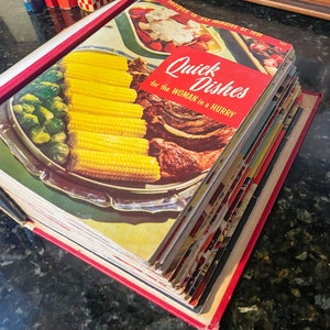 May include: A stack of vintage cookbooks with a red spine and the title "Quick Dishes for the Woman in a Hurry" on the top book. The cover of the top book features a photo of corn on the cob, green beans, and a roast.
