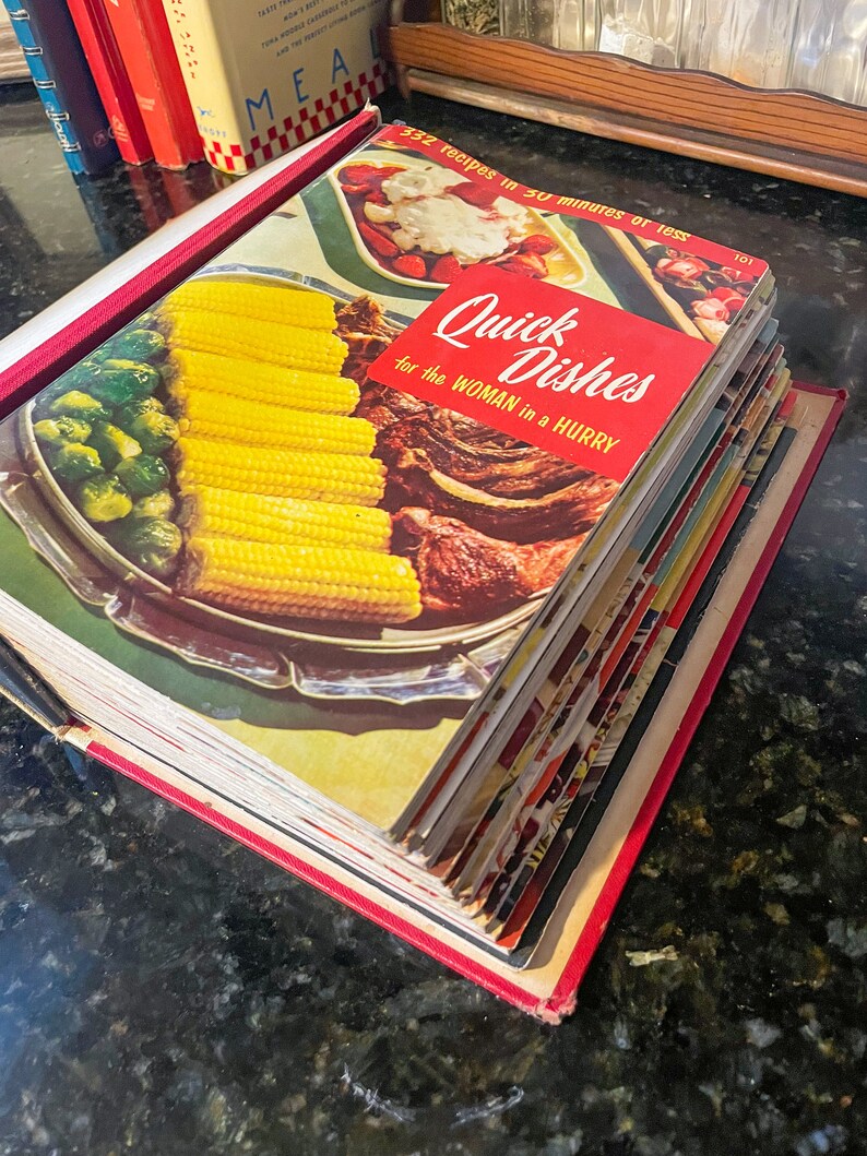 May include: A stack of vintage cookbooks with a red spine and the title "Quick Dishes for the Woman in a Hurry" on the top book. The cover of the top book features a photo of corn on the cob, green beans, and a roast.