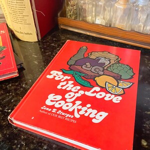 May include: A red hardcover cookbook titled "For the Love of Cooking" by Lena E. Sturges, author of "Our Best Recipes". The cover features a colorful illustration of vegetables, herbs, and a pot of food.