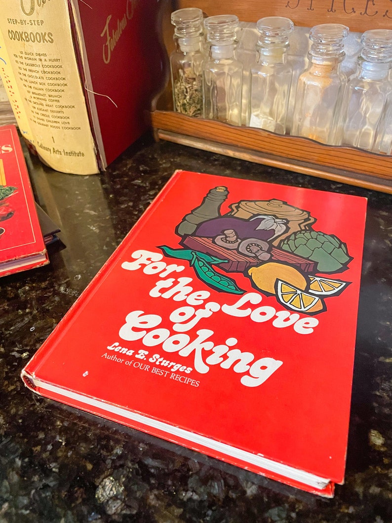 May include: A red hardcover cookbook titled "For the Love of Cooking" by Lena E. Sturges, author of "Our Best Recipes". The cover features a colorful illustration of vegetables, herbs, and a pot of food.