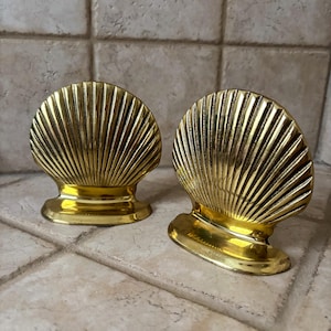 May include: Two gold-colored seashell-shaped decorative objects. Each shell has a detailed, ridged design and sits on a small, rounded base. The objects are displayed on a light-colored, textured surface.