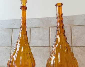 May include: Two amber-colored glass bottles with textured surfaces. One bottle has a tall, pointed stopper with a textured design. The other bottle is open at the top. Both bottles have a teardrop shape and are displayed on a tiled surface.