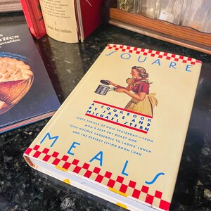 May include: A vintage cookbook titled "Square Meals" by Jane and Michael Stern. The cover features a woman in a red and white dress holding a pot with a red and white checkered pattern.