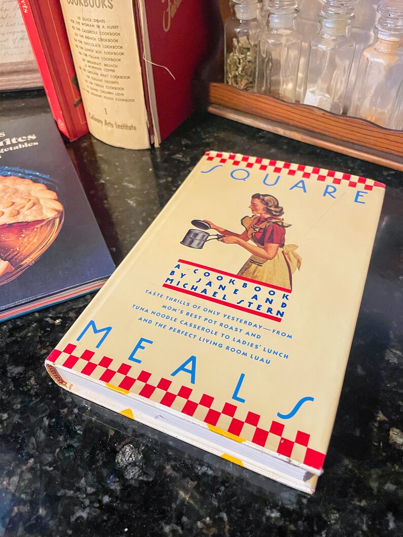 May include: A vintage cookbook titled "Square Meals" by Jane and Michael Stern. The cover features a woman in a red and white dress holding a pot with a red and white checkered pattern.