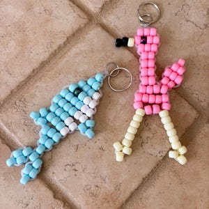 May include: Two handmade keychains crafted from plastic pony beads. One keychain is a light blue and white fish shape, and the other is a pink and cream flamingo shape. Both figures are strung on elastic cord and feature silver metal key rings.