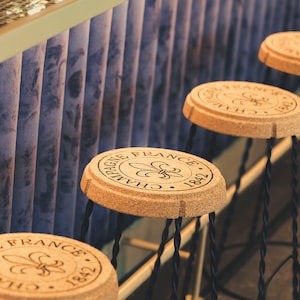Puede incluir: Cuatro taburetes de bar con asientos de corcho, cada uno estampado con "CHAMPAGNE FRANCE 1842" y una flor de lis. Los taburetes tienen patas de metal negro y están dispuestos en fila contra un fondo azul y blanco.