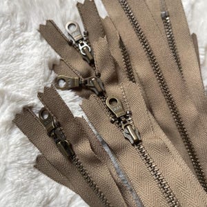 May include: A close-up of several brown zippers with antique brass pulls. The zippers are laid out on a white fuzzy surface.