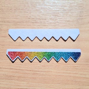 May include: Two decorative, sawtooth-edged felt pieces. One is white, the other features a rainbow glitter design. The pieces are displayed on a light brown wooden surface.