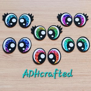 May include: A set of ten colorful googly eyes with black lashes. The eyes are iridescent and have a white pupil. The eyes are arranged in two rows of five. The eyes are on a wooden surface.