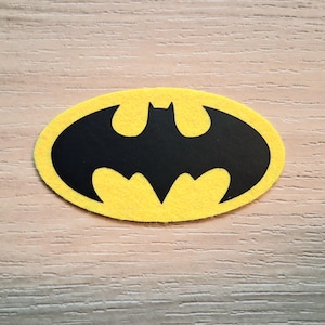 May include: A yellow felt Batman logo with a black bat silhouette in the center. The logo is oval-shaped and sits on a light brown wooden surface. The felt has a slightly textured appearance.