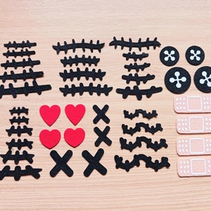 May include: A set of felt craft supplies for creating a monster or spooky character. The set includes black felt stitches, red felt hearts, black felt X's, black felt zigzags, black felt buttons, and beige felt bandages.
