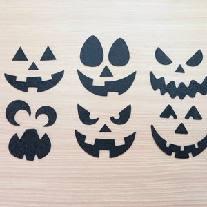 May include: Six black felt Halloween pumpkin face cutouts on a light wood background. The cutouts include a traditional jack-o-lantern face, a scary face with sharp teeth, a face with eyes and a wide mouth, a face with eyes and a small mouth, a face with eyes and a small mouth, and a face with eyes and a small mouth.