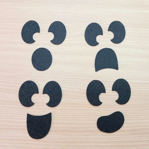 May include: Two sets of black felt ghost face cutouts. Each set includes two eyes, a mouth, and a nose. The eyes are shaped like ovals with a small cutout in the center. The mouths are shaped like a crescent moon. The noses are round.