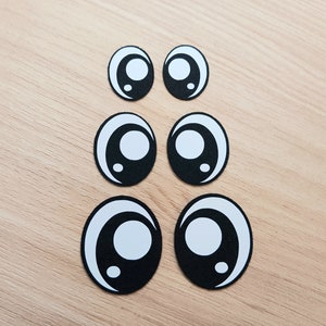 May include: Six black and white googly eyes of different sizes. The eyes are on a light brown wooden surface.