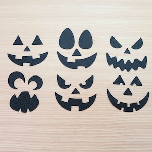 May include: Six black paper cutouts of different jack-o'-lantern faces on a light wood background.