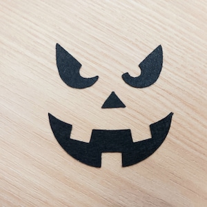 May include: Black paper cutouts of a scary face on a light wood background. The face has two eyes, a nose, and a wide mouth.