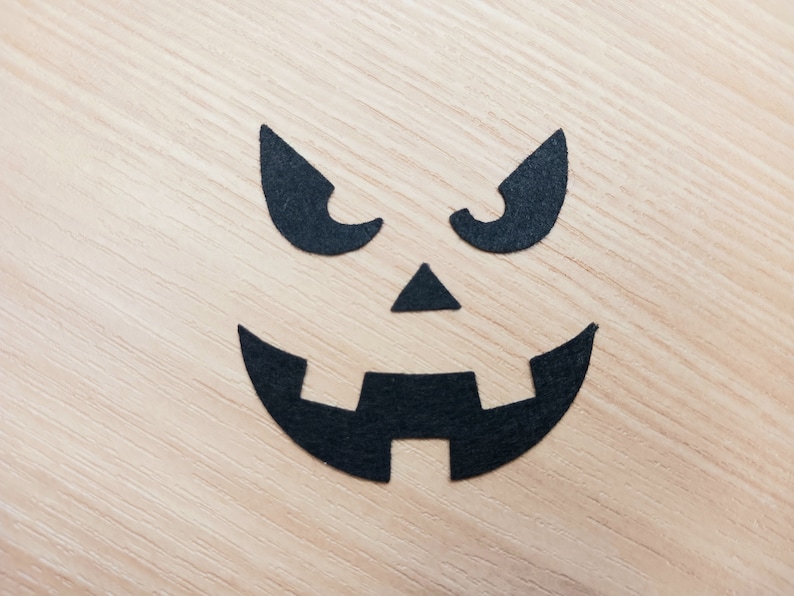 May include: Black paper cutouts of a scary face on a light wood background. The face has two eyes, a nose, and a wide mouth.