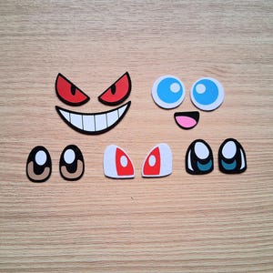 May include: Assortment of cartoon-style face elements, including red angry eyes, a smiling mouth, blue eyes, and various eye pairs. The elements are in different shapes and colors, perfect for crafting or design projects.