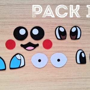 May include: A collection of felt face pieces, including black and white eyes, red cheeks, a pink mouth, and blue and white eye shapes. The text "PACK 1" is displayed in white, above the pieces, on a wooden surface.