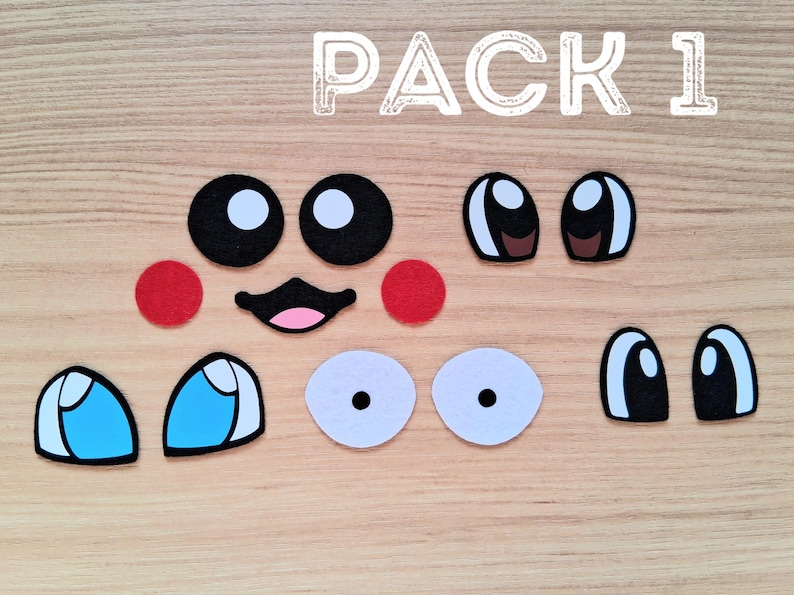May include: A collection of felt face pieces, including black and white eyes, red cheeks, a pink mouth, and blue and white eye shapes. The text "PACK 1" is displayed in white, above the pieces, on a wooden surface.