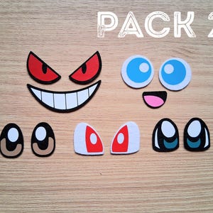 May include: A collection of cartoon face cutouts, including eyes, mouths, and eyebrows. The set features various expressions with colors like red, white, black, blue, and pink. The text "PACK 2" is visible at the top.