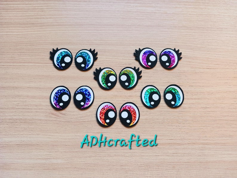 May include: A set of ten colorful googly eyes with black lashes. The eyes are iridescent and have a white pupil. The eyes are arranged in two rows of five. The eyes are on a wooden surface.