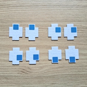 May include: Eight white felt cross shapes with a square of blue felt in the center of each. The shapes are arranged in two rows of four on a light brown wooden surface. The design is reminiscent of pixel art.